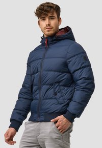 Chaqueta de abrigo azul marino con cremallera frontal, bolsillos laterales y puños acanalados. Presenta una capucha con cordones rojos y diseño acolchado.