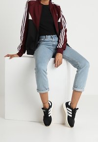 Mörkröd och svart zip-up jacka med vita ränder, i kombination med ljusblå jeans med upprullade fållar och svarta sneakers med vita accenter.