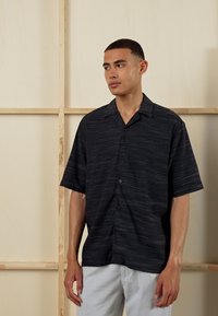 Short-sleeve black shirt with subtle horizontal stripes, button-up front, and a relaxed fit, paired with light-colored pants.