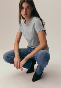 Siva bombažna majica z okroglim izrezom in majhnim zlatim logotipom. Združena z visokim pasom modrih džinsov in črnimi koničastimi gležnjaki.