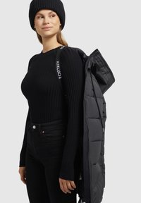 Zwarte geribbelde lange mouwen trui gecombineerd met zwarte skinny jeans, model houdt een zwarte gewatteerde pufferjack met een logo strap vast. Zwarte gebreide beanie.