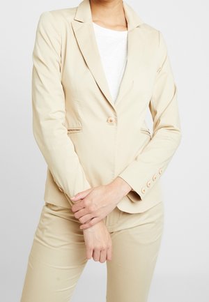 Vrouw in beige colbert en broek met wit overhemd, staand met gevouwen handen voor zich tegen een effen achtergrond.