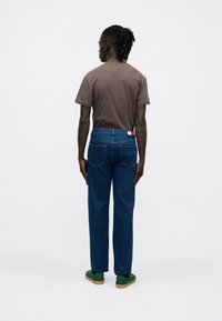 Brūns īsroku t-krekls, zilas džinsa bikses ar taisnu griezumu, sarkans un balts uzraksts uz jostasvietas, zaļas kurpes ar brūnganu zoli.