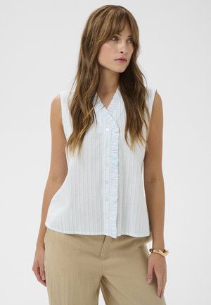 Femme aux longs cheveux bruns portant un chemisier sans manches bleu clair à rayures avec un détail de volants, un pantalon beige et un bracelet en or au poignet droit.