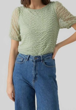 Femme portant un chemisier à manches courtes texturé vert clair associé à un jean en denim bleu taille haute, main sur la hanche.