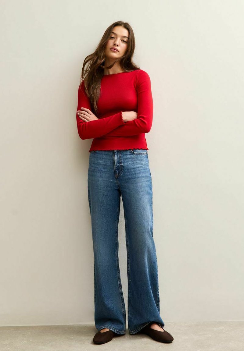 Young woman with long brown hair stands against wall wearing red long-sleeve top, wide-leg blue jeans, and black flats with arms crossed.
