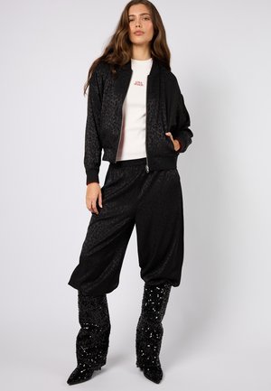 Czarna kurtka bomber z nadrukiem w panterkę, noszona na białej bluzce, zestawiona z luźnymi czarnymi spodniami i błyszczącymi, sięgającymi do kolan kozaki.