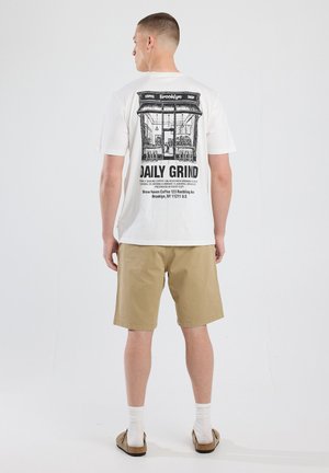 White cotton t-shirt with a black graphic print featuring a coffee shop scene and text, paired with khaki shorts and slip-on sandals.