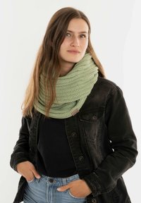 Lichtgroene gebreide infinity-sjaal met een gestructureerd patroon, gedragen over een zwarte top en een zwarte spijkerjack. Casual look met handen in de zakken.
