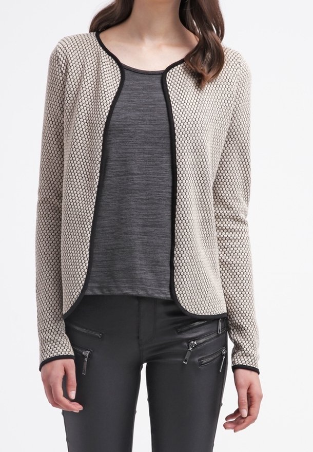 Beige strukturovaný cardigan s černým lemem, vrstvený přes tmavě šedý top. V kombinaci s černými kalhotami s zipovými detaily.