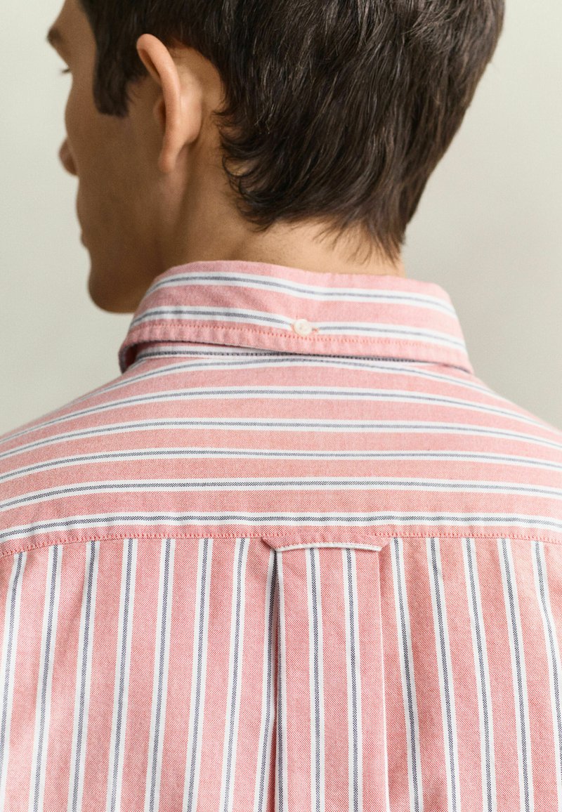 Vue de dos d'une personne portant une chemise à rayures roses et blanches avec les cheveux courts et foncés, sur un fond uni.