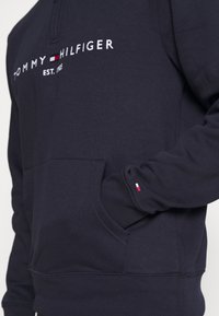 Felpa blu navy con tasca frontale, caratterizzata dal logo bianco "TOMMY HILFIGER" e dettagli rossi, bianchi e blu sul polsino delle maniche.