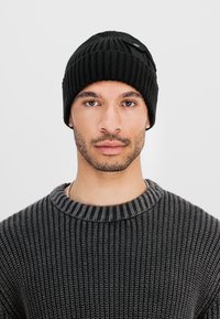 Man with trimmed beard wearing a black knit beanie and dark gray ribbed sweater, facing forward against a plain light background.