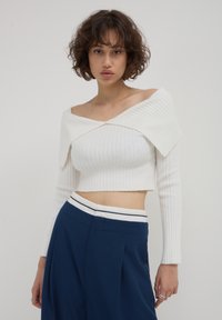 Femme jeune aux cheveux courts et bouclés porte un pull court blanc à épaules dénudées et un pantalon taille haute bleu marine avec une ceinture blanche, se tenant devant un fond uni.