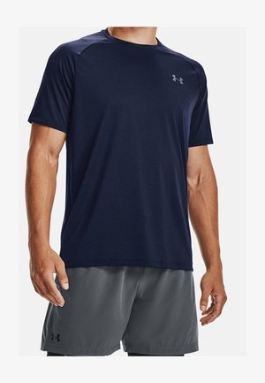 Homme portant un maillot de sport bleu marine à manches courtes et un short de sport gris, debout avec les bras détendus le long du corps, fond non visible.