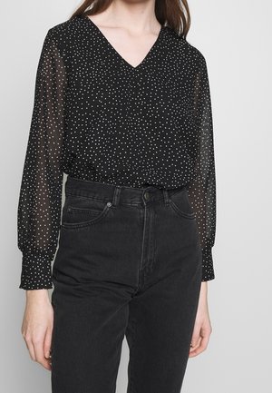 Schwarze Bluse mit langen Ärmeln und V-Ausschnitt, mit weißen Punkten, aus einem transparenten Material. Kombiniert mit hoch taillierten schwarzen Jeans.