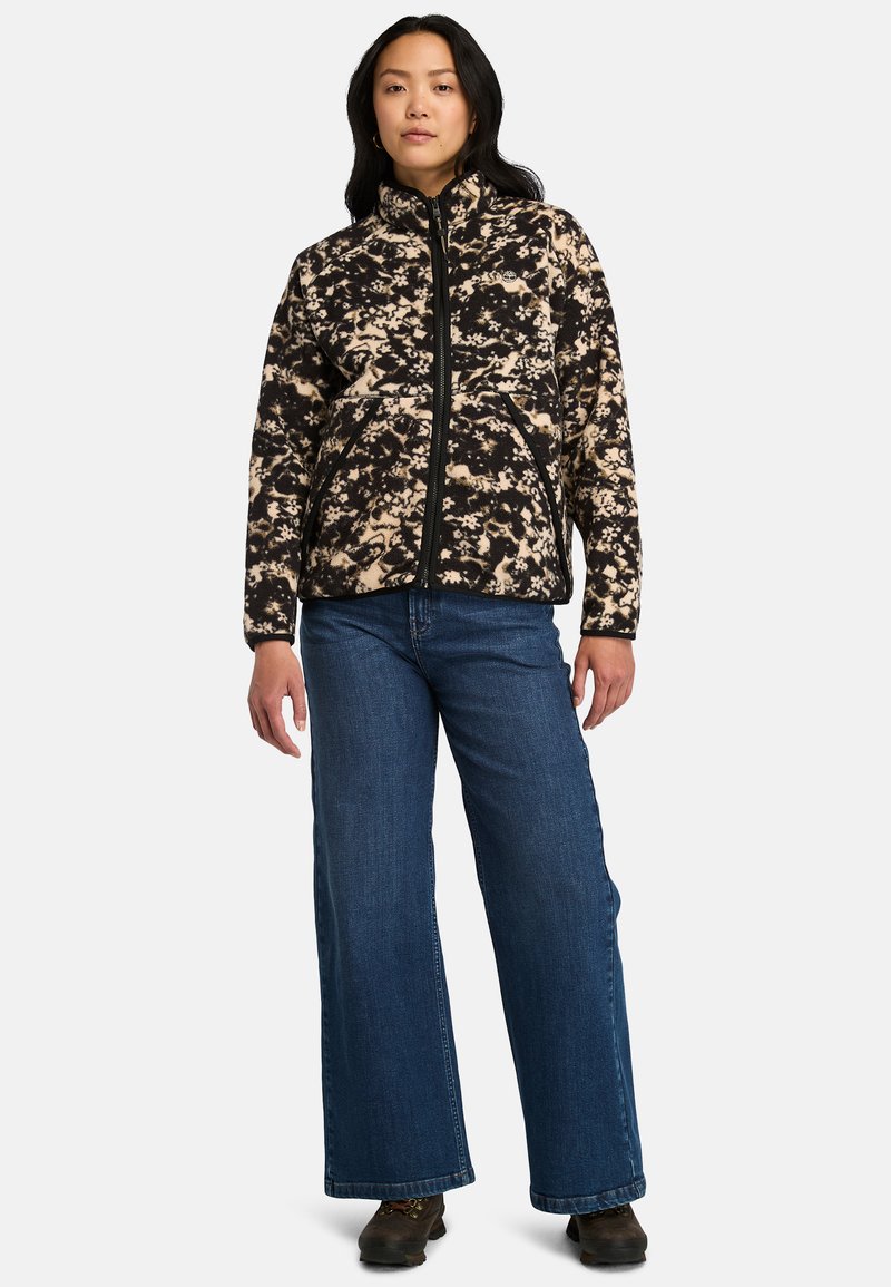 Fleecejacka i svart och tan abstrakt mönster, hel dragkedja, hög krage, framfickor, kombinerad med blå vida jeans och bruna skor.