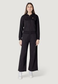 Felpa nera con zip e coulisse, con dettagli del logo; abbinata a pantaloni neri a gamba larga; indossata con sneakers bianche. Tessuto liscio e vestibilità comoda.