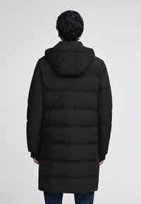 Svart vadderad lång kappa med huva, med en kviltad design och mjuk textur. Kappan når till mitten av låret och har långa ärmar.