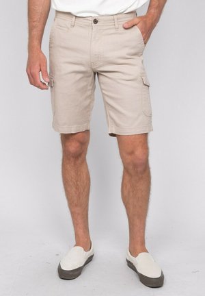 Homme portant un short cargo beige clair, une chemise blanche et des chaussures blanches sans lacets, une main dans la poche, debout sur un fond uni.