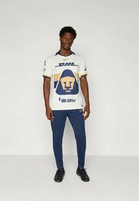 Biela futbalová dres s modrými a zlatými akcentmi, so znakom leva a brandingom "DHL", skombinovaný s námorníckymi teplákmi a čiernymi teniskami.