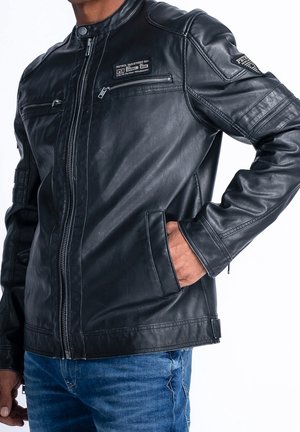 Homme portant une veste en cuir noire avec une poche poitrine zippée et une main dans la poche latérale, associée à un jean bleu, sur fond blanc.