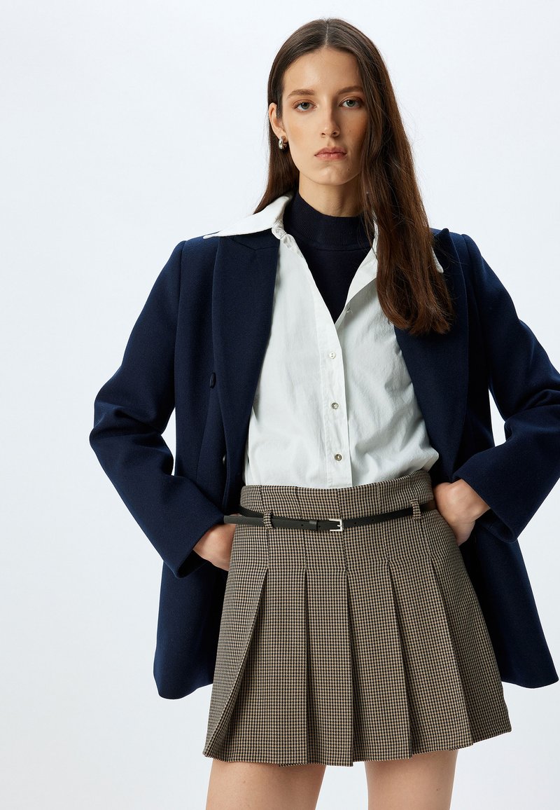 Blazer navy, camicia bianca a bottoni, dolcevita nera e gonna plissettata a quadretti con cintura nera, che mette in mostra il design strutturato e le texture.