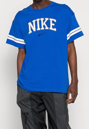 Blå bomuld T-shirt med stort hvidt "NIKE"-logo og sort swoosh. Korte ærmer med hvide striber på ærmerne. Casual pasform.