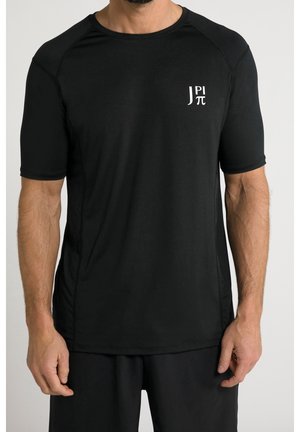Mężczyzna w prostym czarnym t-shirtcie z krótkim rękawem, z małym białym logo "J PI π" na lewej piersi na jasnym tle.