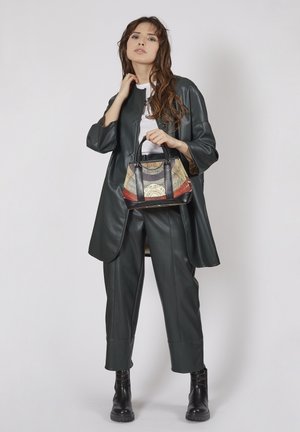 Frau in dunkelgrüner Lederjacke und Hose, die eine gemusterte Handtasche hält und vor einem schlichten Hintergrund in heller Farbe steht.