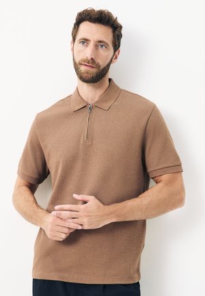 Bărbat cu barbă, purtând un tricou polo maro cu mâneci scurte și fermoar frontal, stând în picioare pe fundal alb, cu mâinile încleștate aproape de talie.