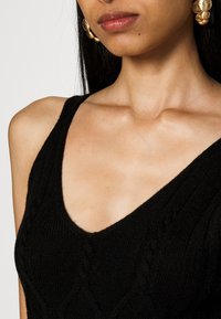 Black knitted tank top featuring a V-neck design with textured cable patterns, paired with large gold hoop earrings.