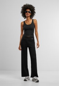 Schwarzes Tanktop und weite Hose aus weichem Stoff. Das Outfit wird mit grauen Sportschuhen und dunklen Sonnenbrillen kombiniert.