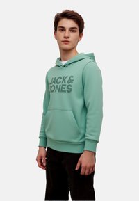 Hoodie verde menta con tasca frontale, coulisse regolabile e grande logo stampato "JACK & JONES" in verde scuro. Texture liscia.