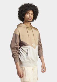 Ung man med lockigt hår, iklädd en beige, brun och gräddfärgad färgblockerad hoodie samt ljusa cargobyxor, stående framför en vit bakgrund.