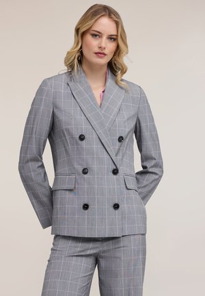 Joven mujer con un traje a cuadros gris de doble botonadura con botones negros y pantalones a juego, posando ante un fondo neutro.