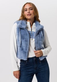 Hellblaues Kunstpelz-Weste mit Kapuze, Reißverschluss und Seitentaschen, getragen über einem weißen Langarmshirt und dunkelblauen Jeans.