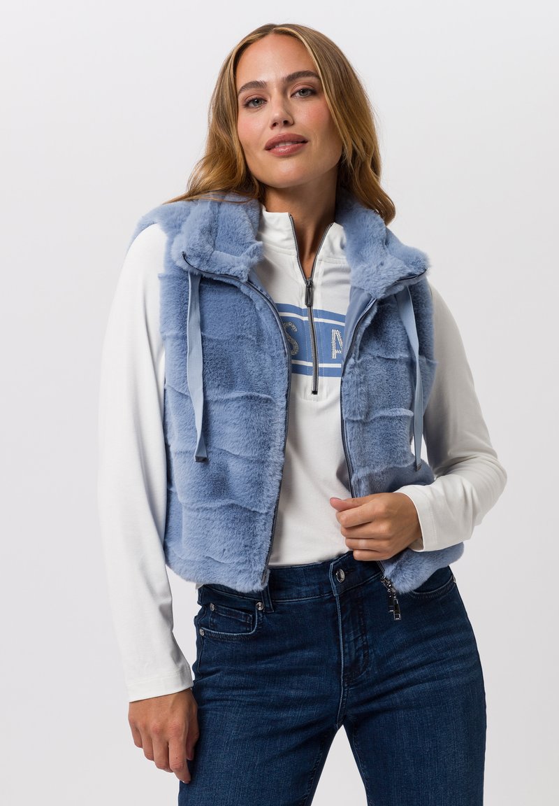 Hellblaues Kunstpelz-Weste mit Kapuze, Reißverschluss und Seitentaschen, getragen über einem weißen Langarmshirt und dunkelblauen Jeans.