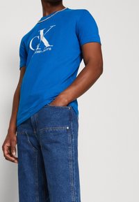 Modrá bavlněná trička s velkým bílým logem CK, spárovaná s tmavě modrými džíny. Džíny mají viditelné švy a kapsy.