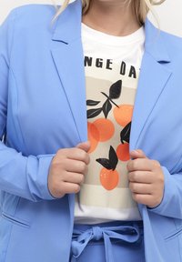 Blazer bleu clair en tissu lisse, porté par-dessus une chemise crème avec un graphisme coloré orange et papillon. Détail de ceinture à la taille.