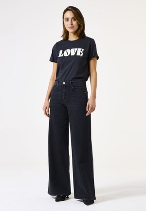 Woman wearing a black T-shirt with "LOVE" text and high-waist wide-leg black jeans, standing against a plain light background.