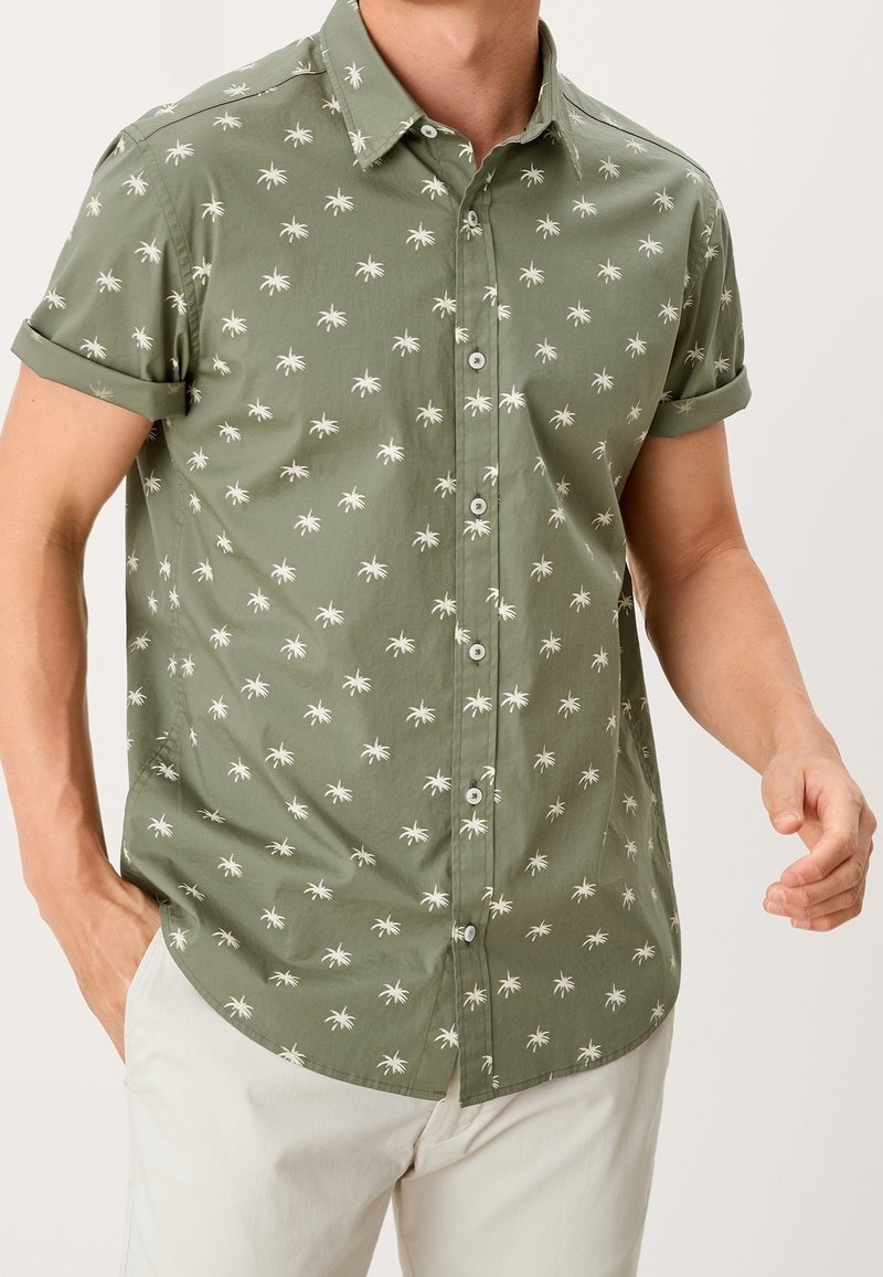Camisa de botones de manga corta en tejido verde oliva, con un patrón de palmeras blancas. Cuello, puños enrollados y un dobladillo curvado.