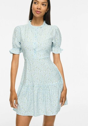 Femme portant une robe florale bleu clair avec des manches bouffantes, un col à volants et des boutons sur le devant, regardant légèrement sur le côté.