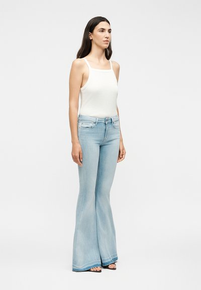 Γυναίκα που στέκεται, φοράει λευκό αμάνικο τοπ και ανοιχτό μπλε ψηλόμεσα καμπάνες τζιν με μαύρα πέδιλα ανοιχτής μύτης σε μονόχρωμο φόντο.