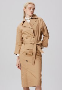 Manteau beige avec une taille ceinturée, boutons double boutonnage, large col et manches longues, associé à une jupe arrivant au genou. Texture de tissu lisse.