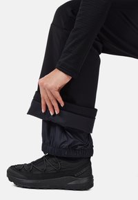Czarne wodoodporne spodnie z elastycznymi mankietami, nałożone na czarny sneaker z teksturowanym materiałem i gumową podeszwą. Ręka poprawia dół.