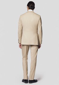 Beżowy garnitur slim fit z pojedynczym rozcięciem z tyłu, wyraźnymi ramionami i czarnymi butami wizytowymi. Tkanina wydaje się gładka i ma dopasowane wykończenie.