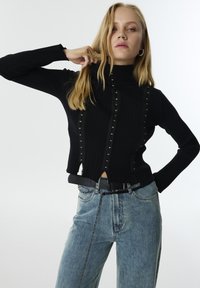 Haut noir à manches longues côtelé avec un col haut, orné de clous en argent le long des coutures avant. Associé à un jean en denim bleu et une ceinture noire.