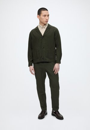 Man staat, draagt een donkergroene, dichtgeknoopte jas en broek, beige overhemd en donkerbruine schoenen, kijkt naar de zijkant.