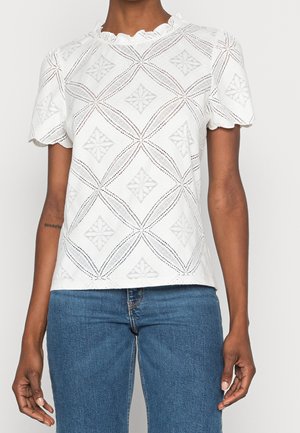 Biała bluzka z krótkim rękawem z ząbkowanymi krawędziami i geometrycznym wzorem z koronki, noszona z niebieskimi dżinsami, prezentująca górną część tułowia i ramiona.